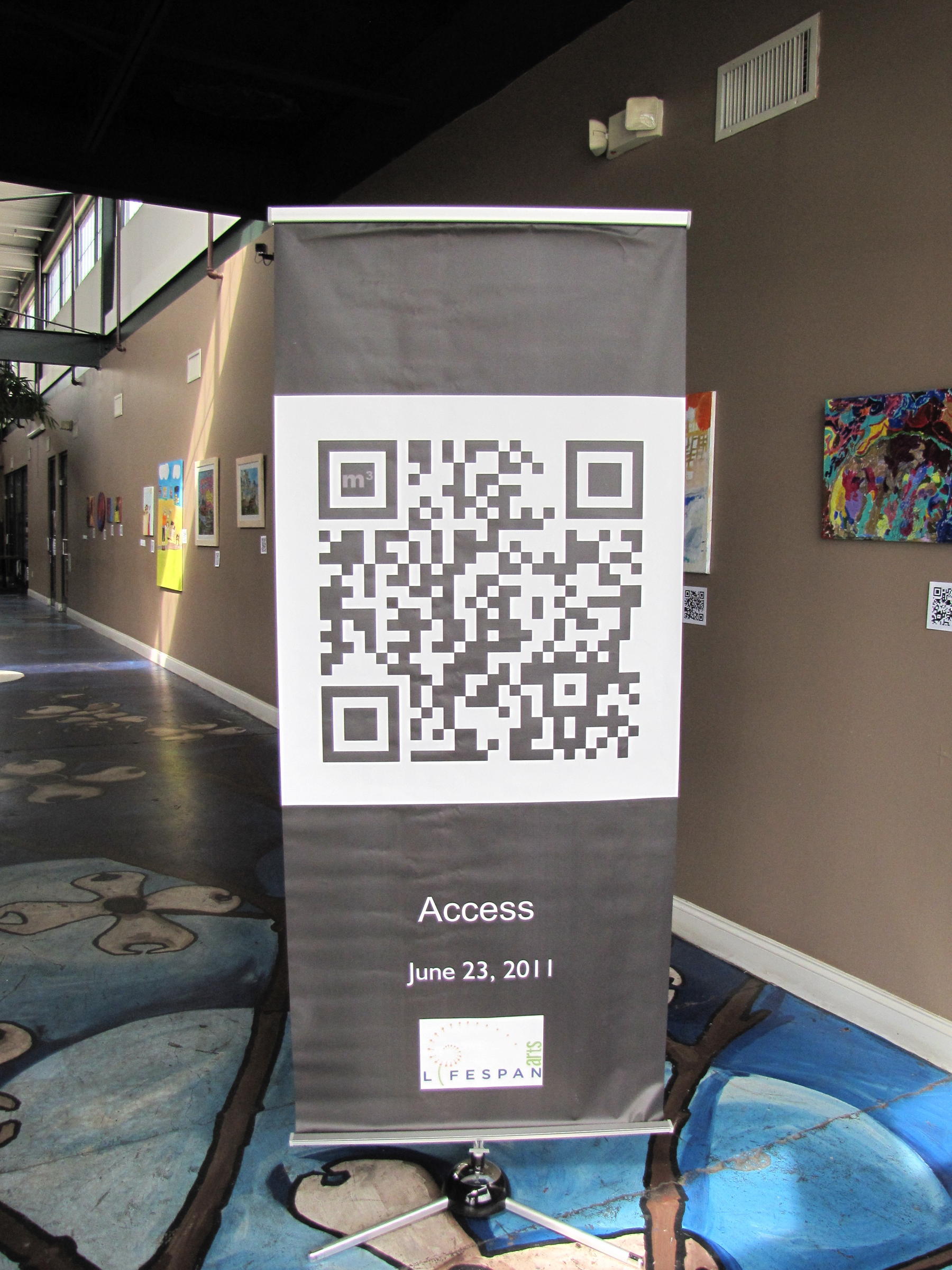 A tall banner featuring a large QR code stands in the entrance of a Charlotte art gallery, surrounded by colorful paintings on the walls. The banner reads Access, June 23, 2011, with the Lifespan Arts logo.
