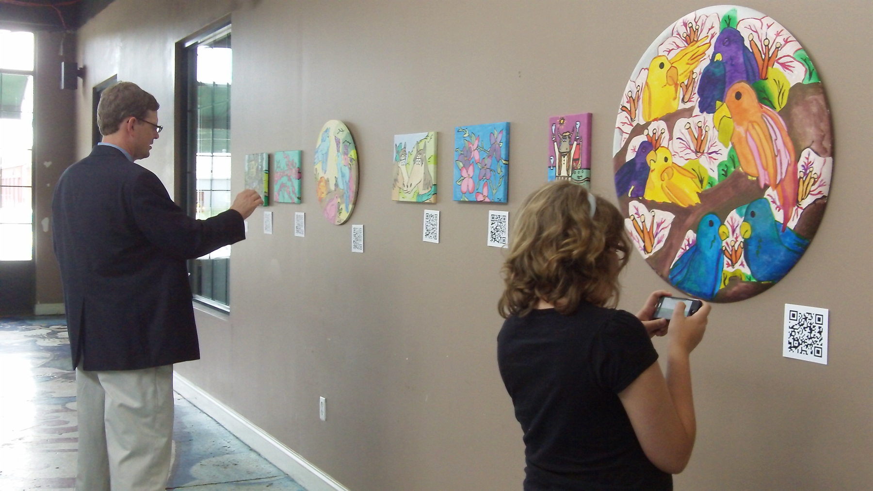 Two gallery visitors examining paintings on a wall. One person holds their phone up to scan a small QR code placed beside a large colorful painting of birds. Other QR codes are visible next to each artwork.